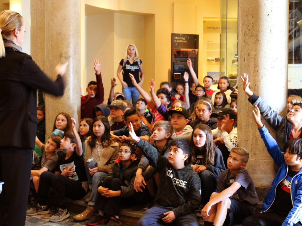 An attentive 5th grade class listens to a Museum Guide, and several children eagerly raise their hands.