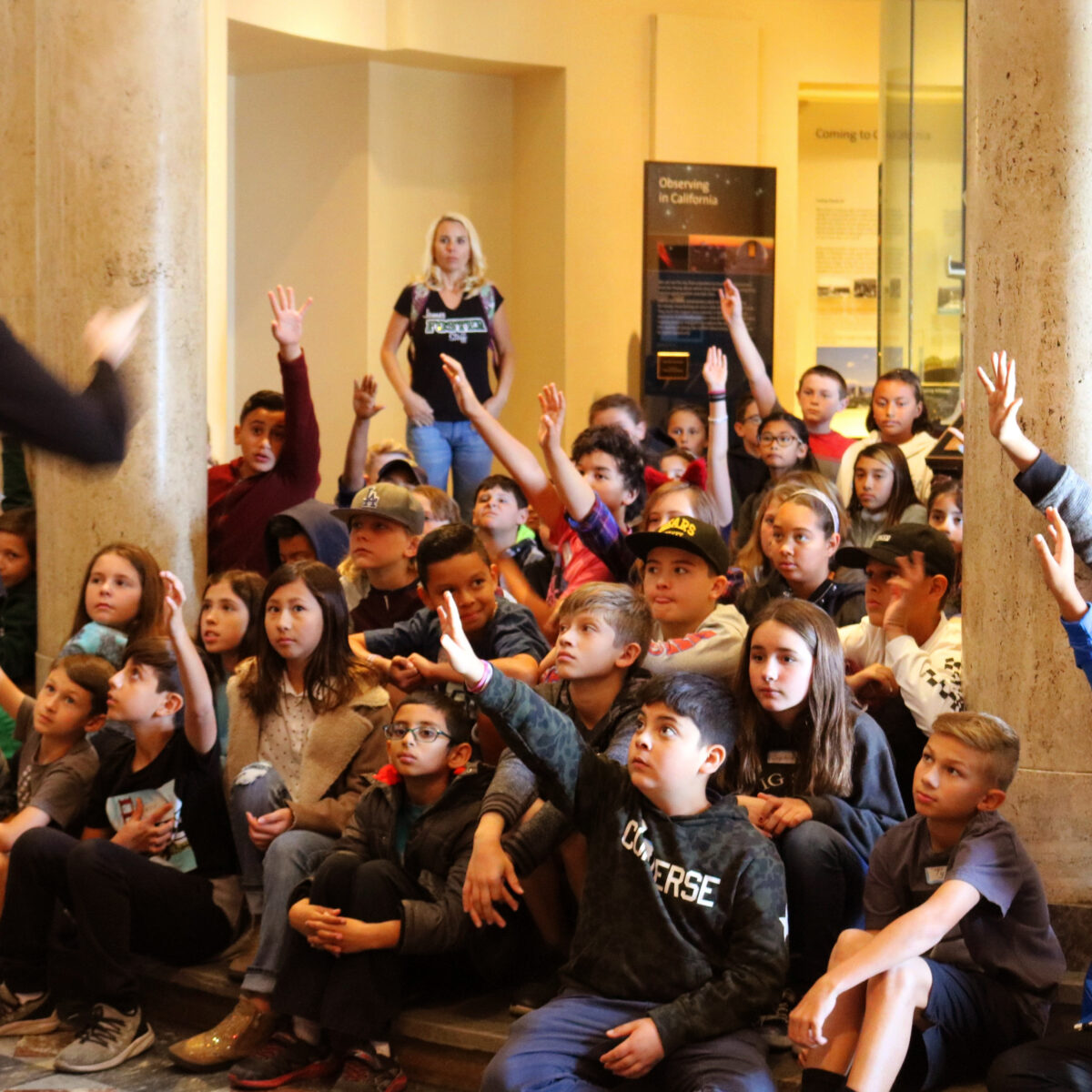 An attentive 5th grade class listens to a Museum Guide, and several children eagerly raise their hands.