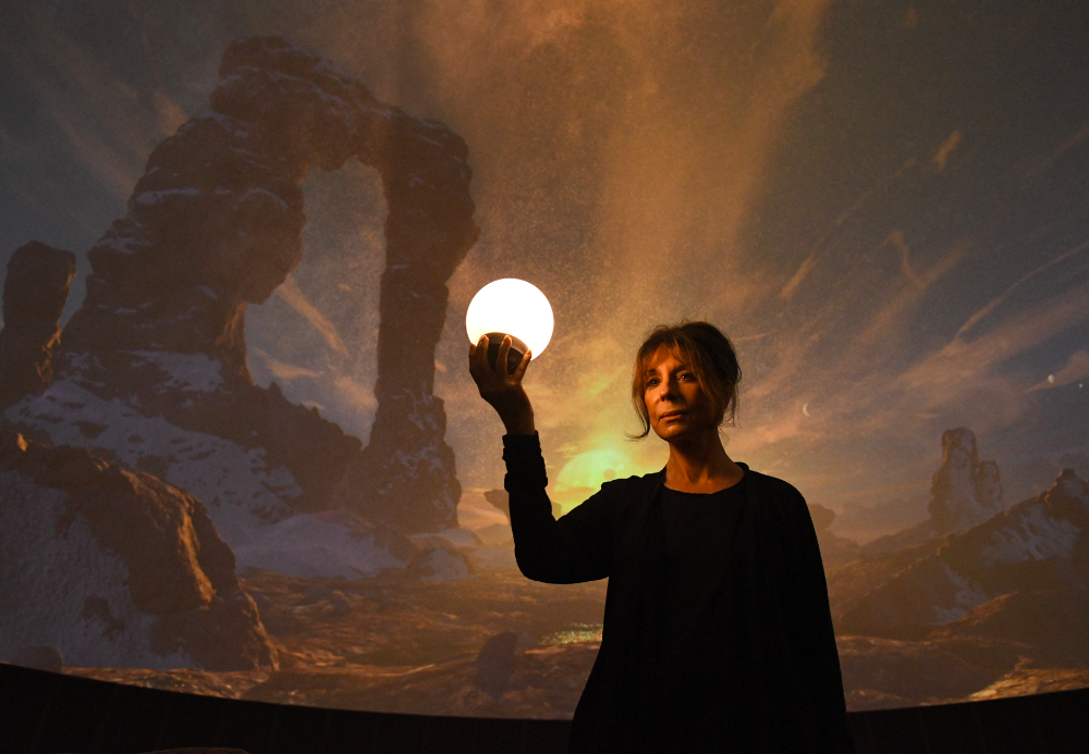 During a preview performance of Signs of Life, Lecturer Suzanne Ford stands in front of a conception of what the surface of an extrasolar planet (Trappist-1) might look like.