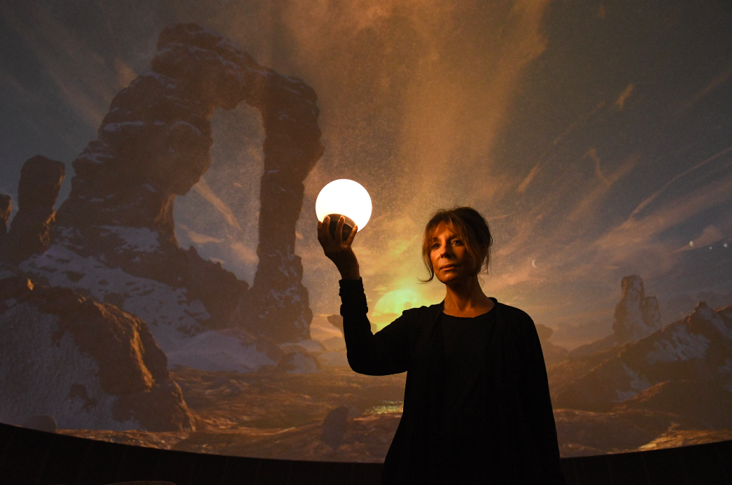 During a preview performance of Signs of Life, Lecturer Suzanne Ford stands in front of a conception of what the surface of an extrasolar planet (Trappist-1) might look like.