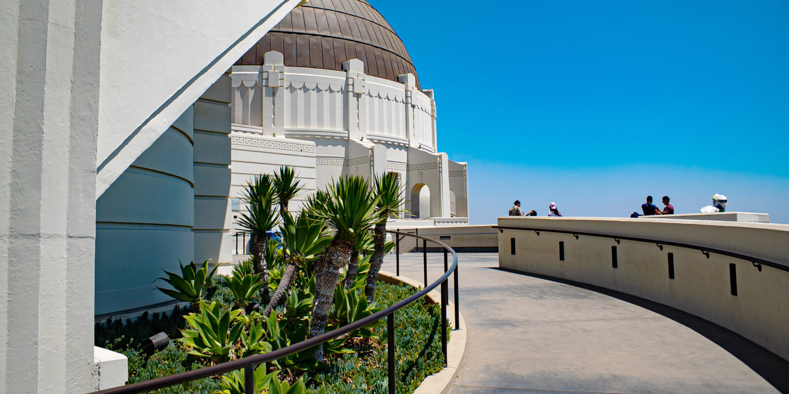 Curved path leading to a white Observatory dome, surrounded by lush green plants. People are enjoying a sunny day against a clear blue sky.